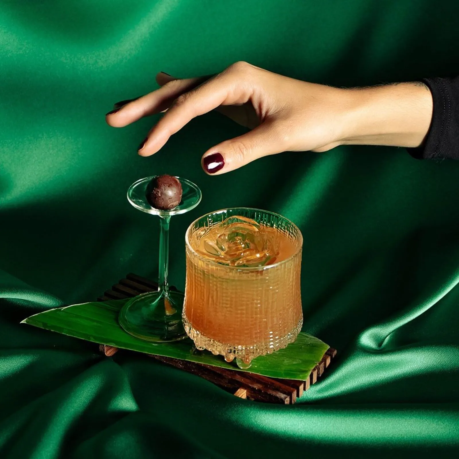 Cóctel elegante acompañado de un bombón de chocolate, presentado sobre una hoja verde con fondo de tela satinada en tono verde. Estilo sofisticado y lujoso en Madrid Cocktail Week