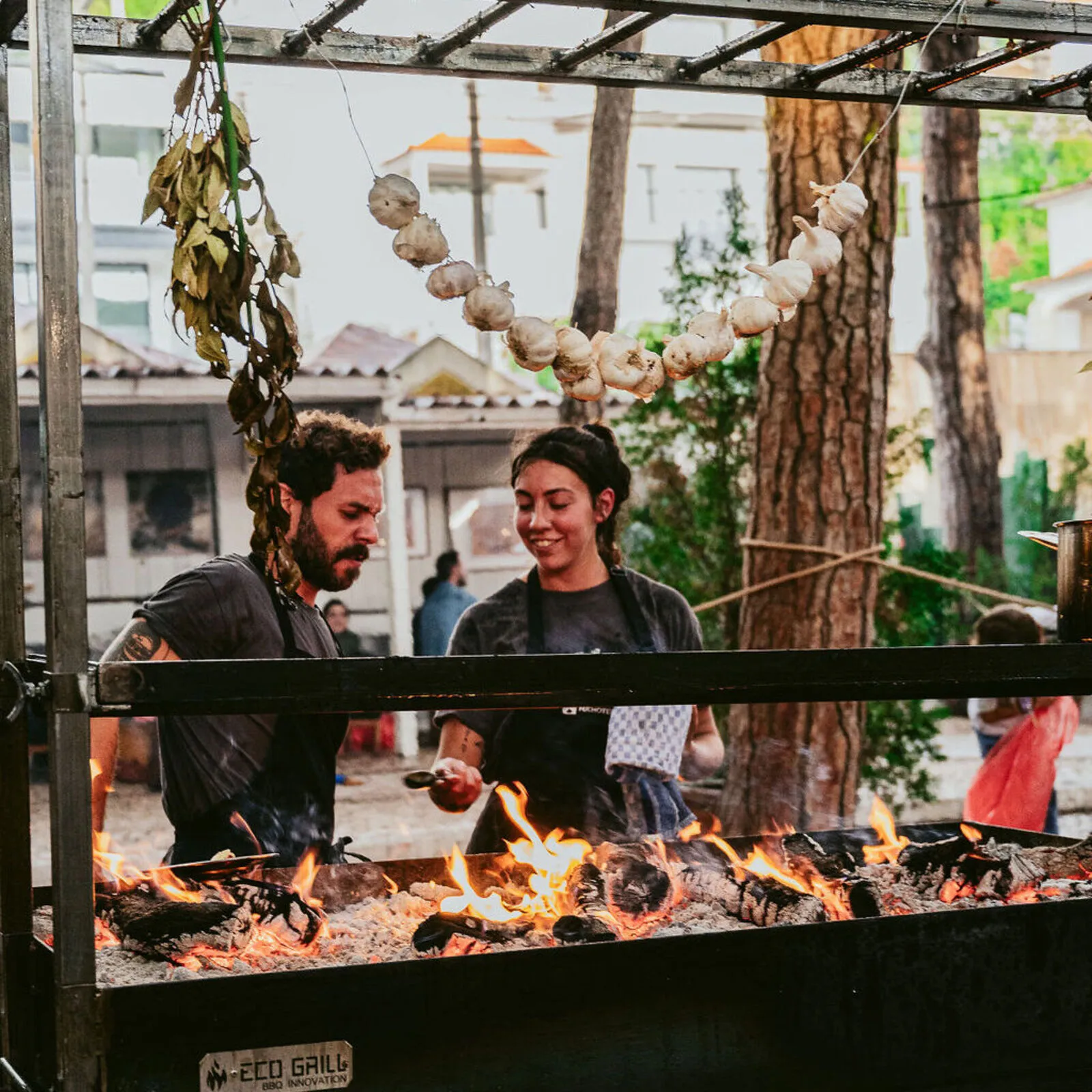 Dos chefs cocinando a la parrilla al aire libre sobre fuego de leña, rodeados de vegetación y adornos colgantes de ajo