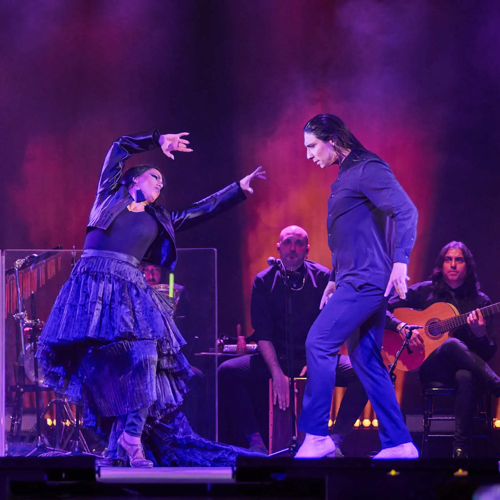 Bailarines de flamenco en un espectáculo de flamenco en Madrid, acompañados de música en vivo con guitarristas y percusionistas, bajo una iluminación dramática en tonos morados y naranjas