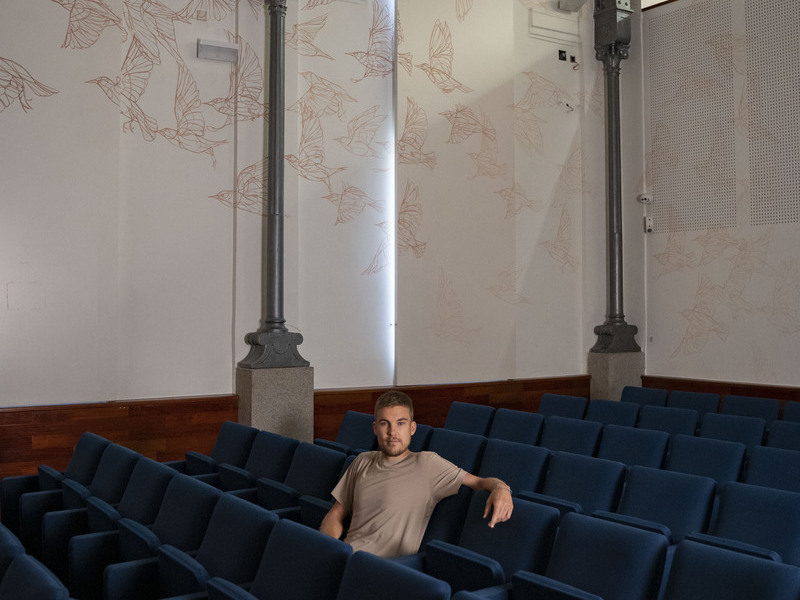 Taquen artista y muralista español sentado en una sala de conferencias decorada con ilustraciones de aves, con asientos azul oscuro y una atmósfera tranquila y artística