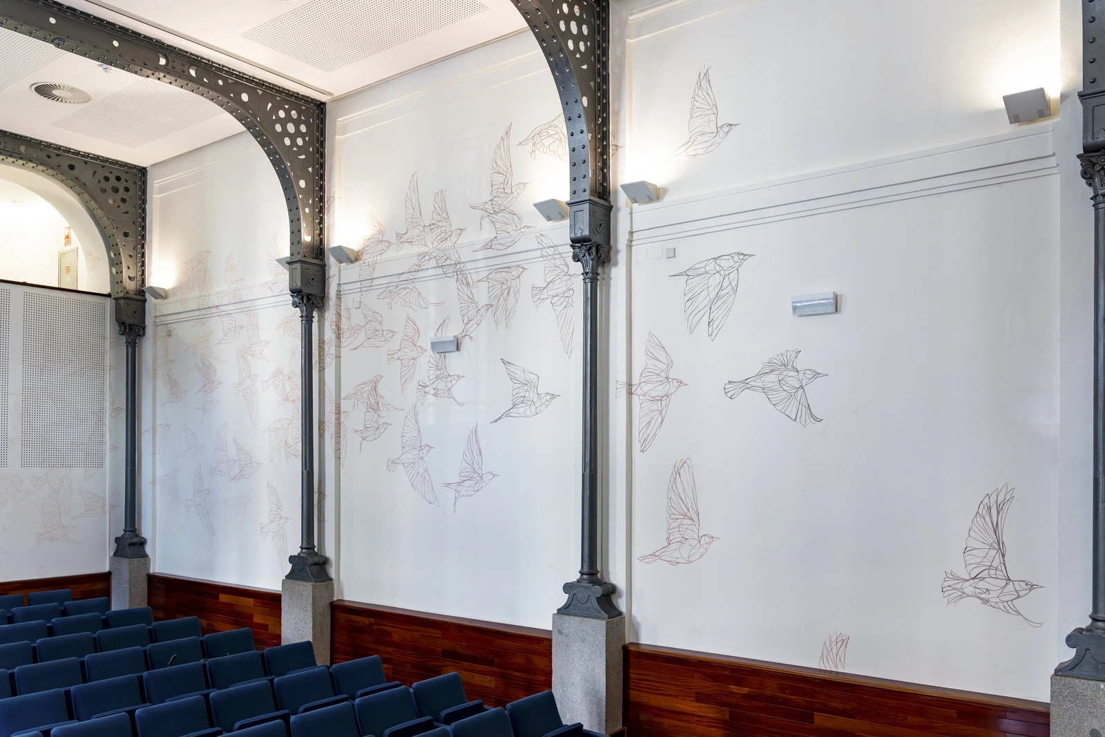 Sala de conferencias decorada con ilustraciones de aves en vuelo sobre paredes blancas, hechas por el Taquen artista y muralista español, con asientos azul oscuro y detalles arquitectónicos industriales