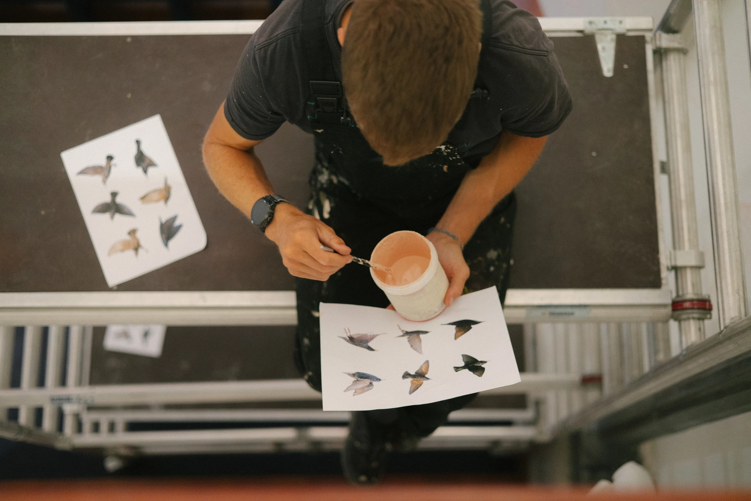 Taquen Artista y muralista español pintando bocetos de aves en una mesa alta, sosteniendo un pincel y un tarro de pintura rosa. Proceso creativo capturado en un entorno artístico y detallista