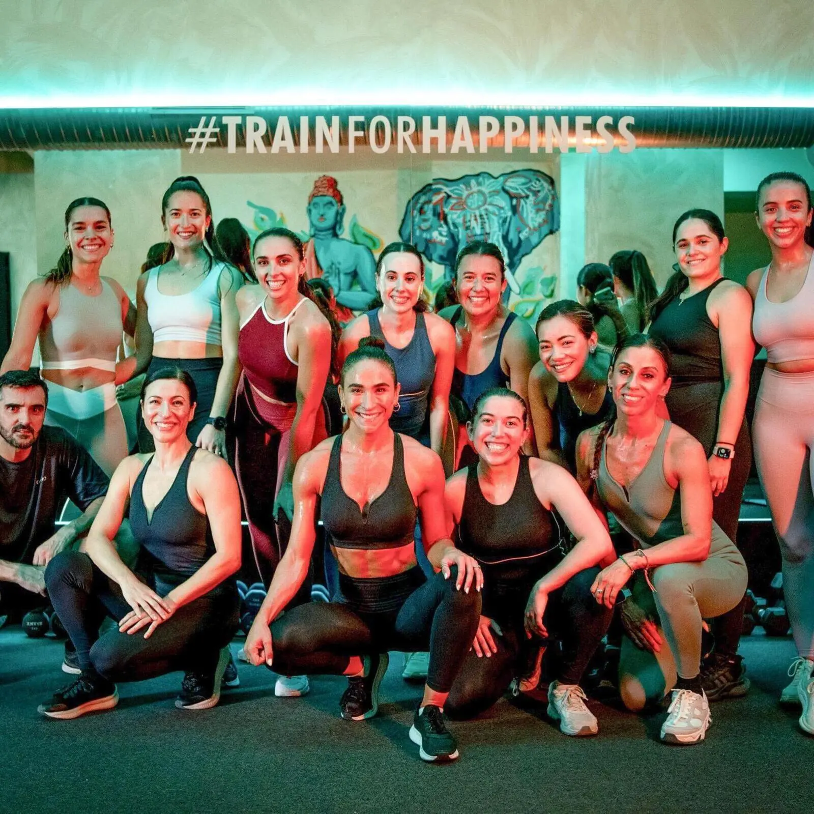 Grupo de personas sonrientes en ropa deportiva posando en un gimnasio después de una sesión de entrenamiento, con el letrero '#TRAINFORHAPPINESS' al fondo y un mural colorido de un elefante. Ambiente iluminado con luces cálidas en tonos rojos y verdes